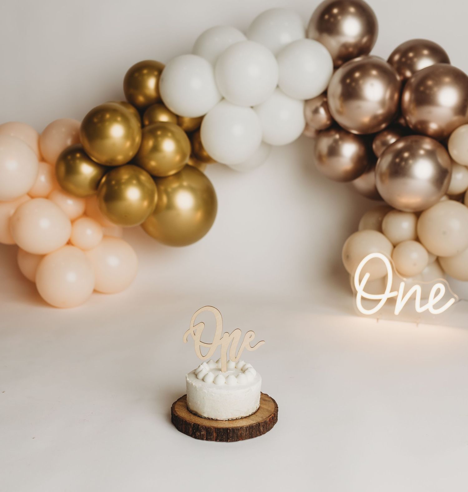 Elegant balloon arch in gold, white and rose gold with 'One' cake topper on wooden slice.