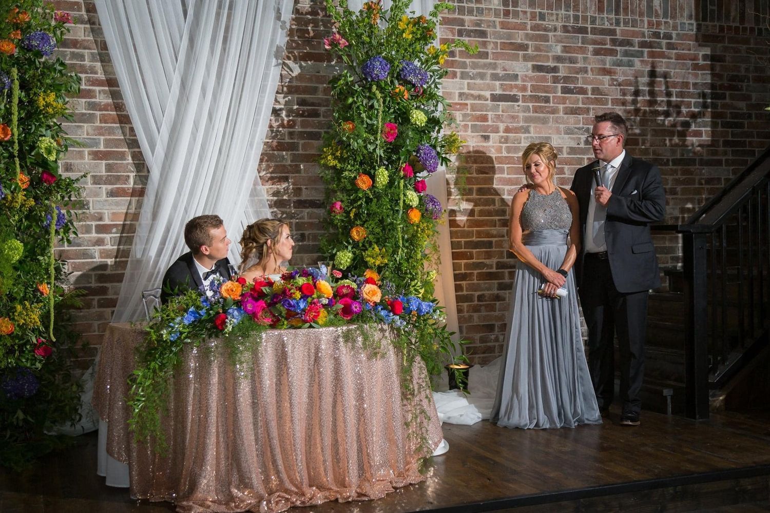 Wedding reception setup with floral arch and sequin tablecloth against exposed brick wall.