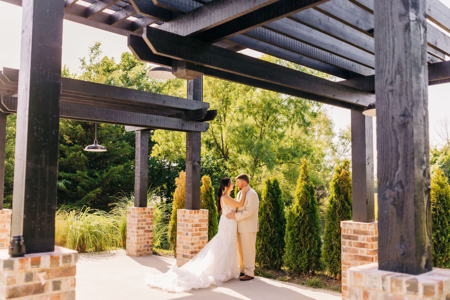 Couple shares a romantic moment under a rustic wooden pergola with brick columns and greenery.