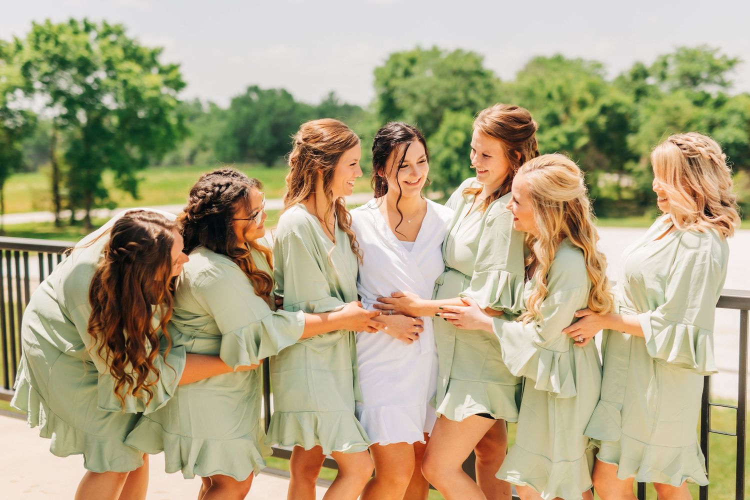 Bridesmaids in matching mint green robes gather in a circle sharing a joyful moment outdoors.