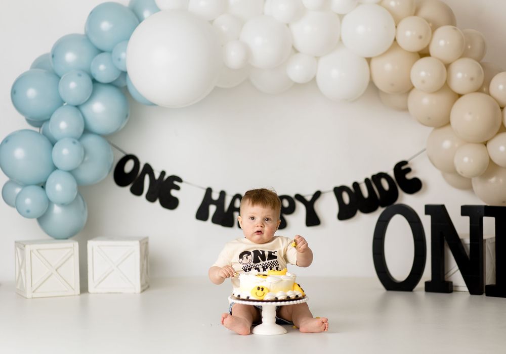 Baby with cake under balloon arch and ONE text decoration for first birthday.