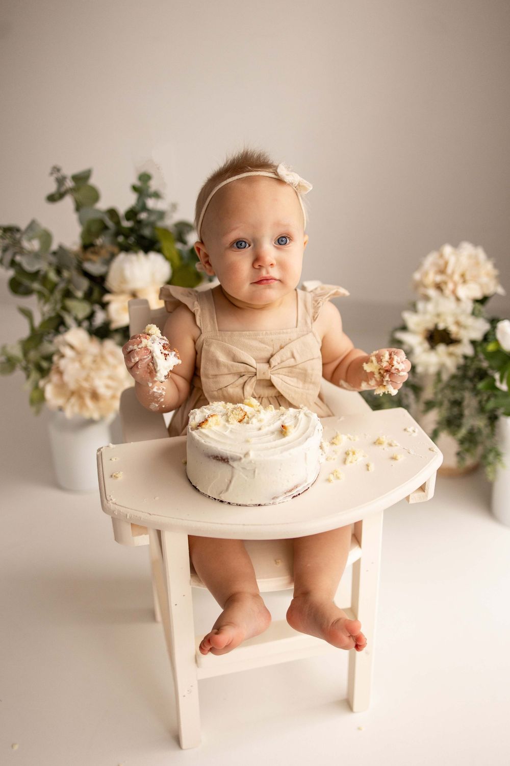 Baby sits in white wooden high chair surrounded by delicate white flowers for cake smash photo session.