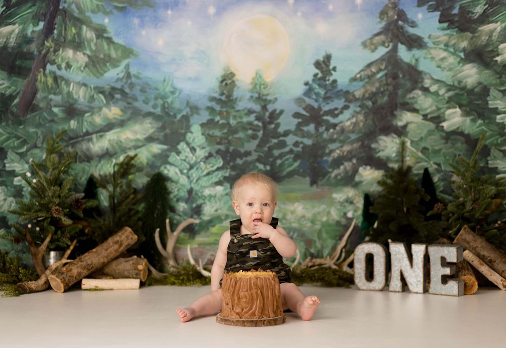 Baby in brown outfit against forest backdrop with ONE sign and woodland decorations.