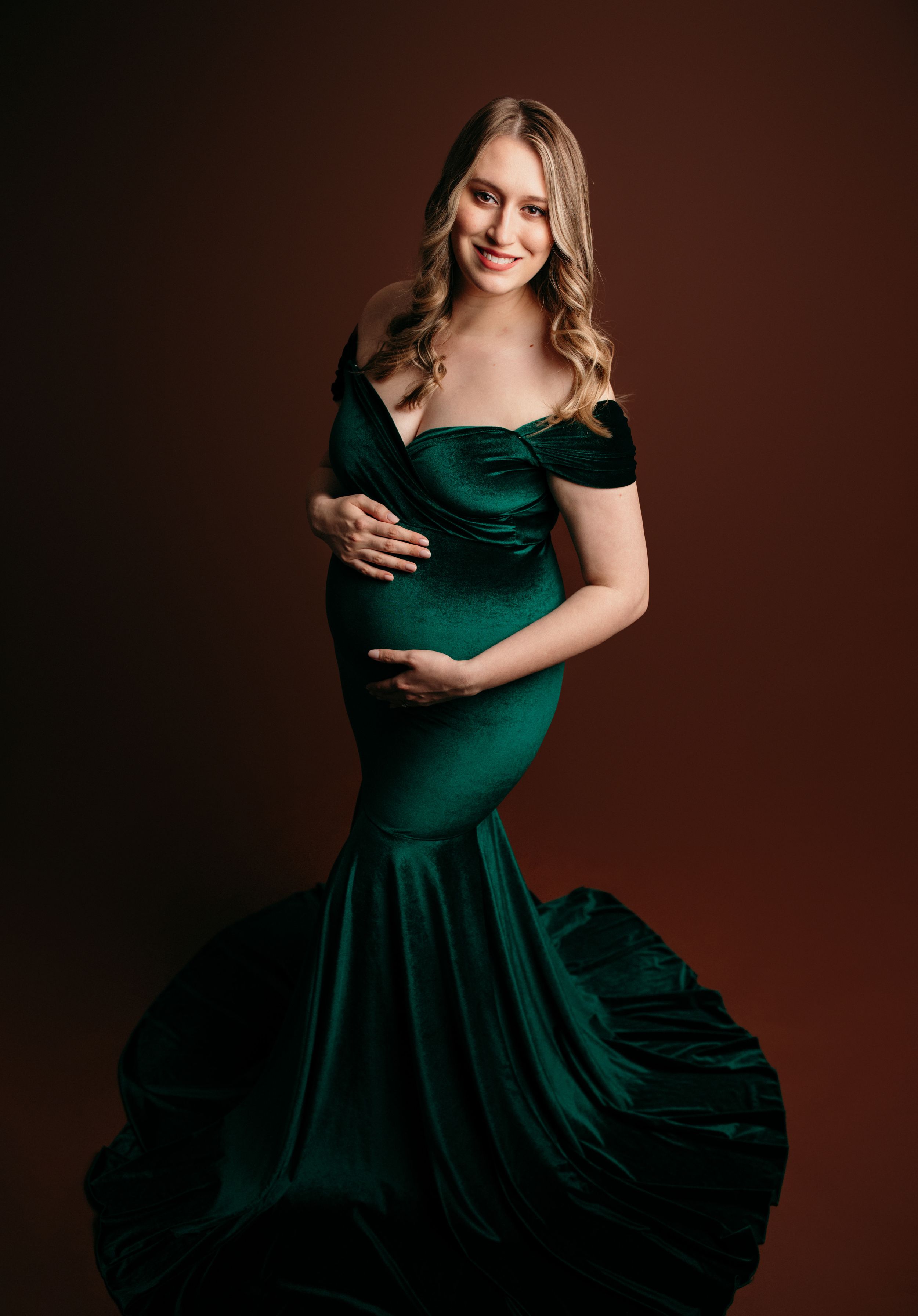 Elegant emerald green velvet maternity gown with sweetheart neckline and flowing skirt against dark background.
