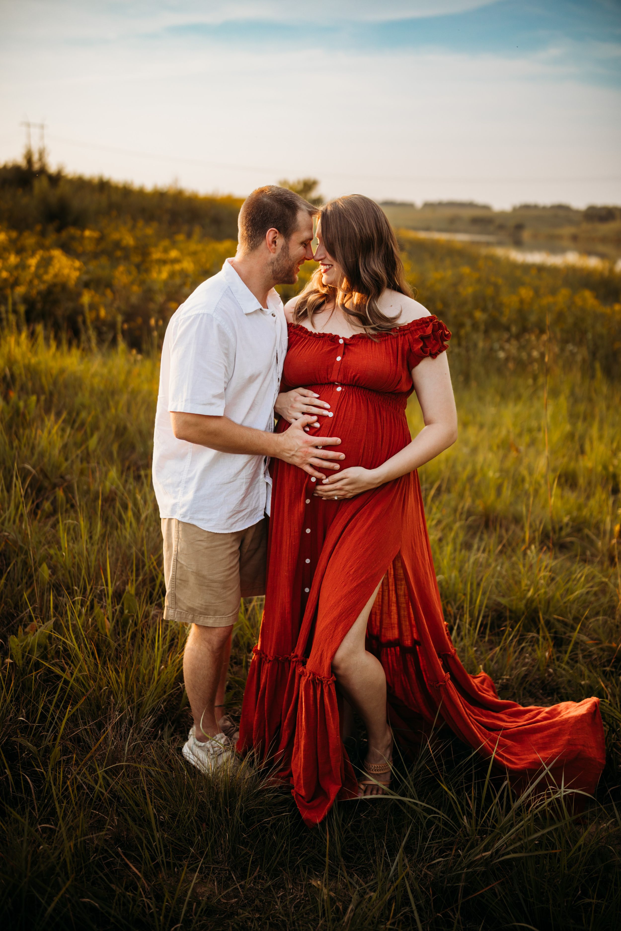 Couple in a field at sunset with flowing red dress and white shirt creating romantic maternity portraits.