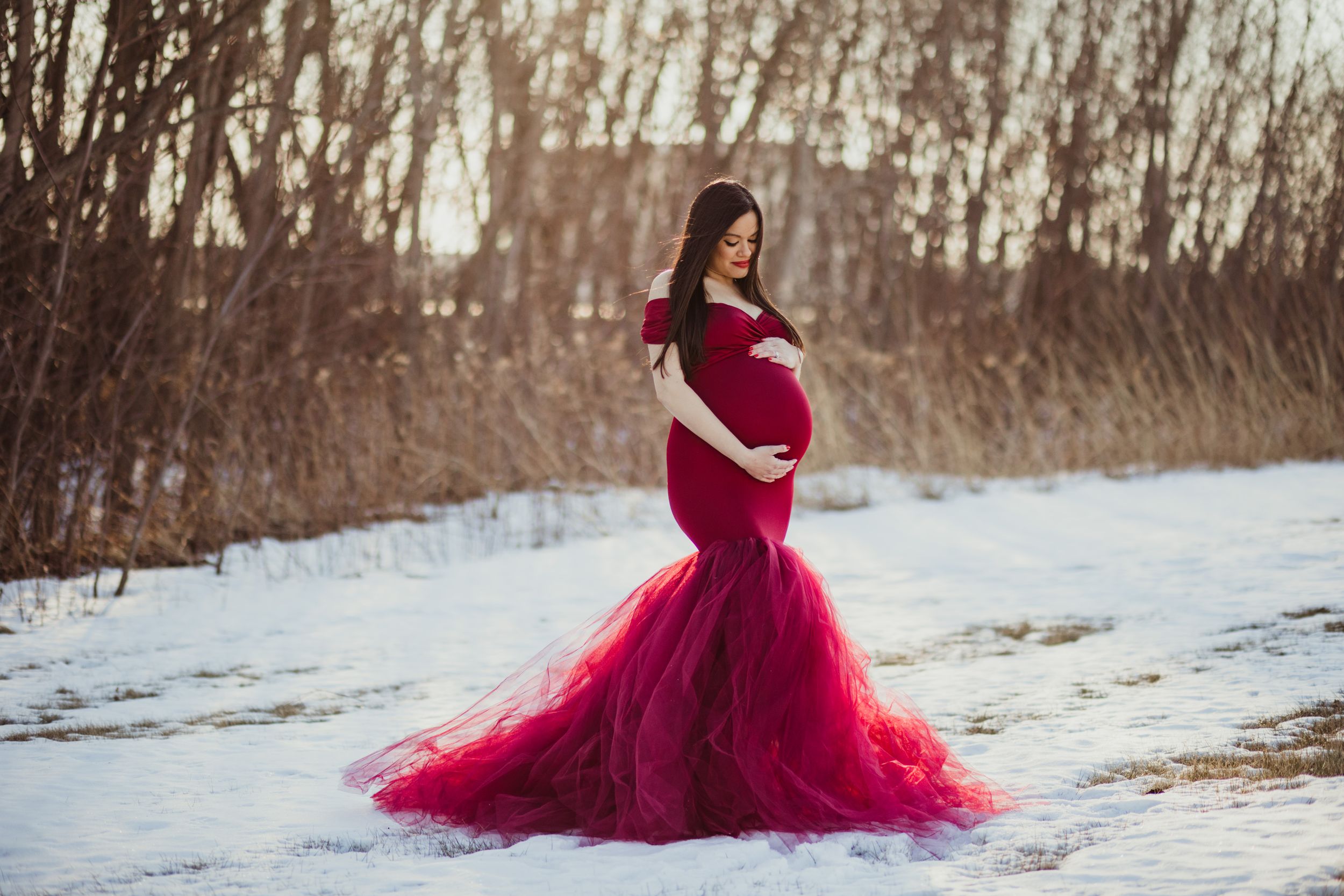A figure in a flowing burgundy gown poses gracefully in a snowy winter landscape at sunset.