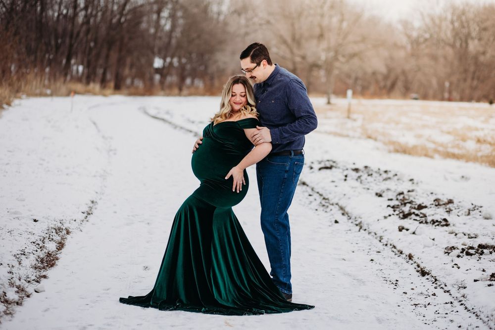 A couple embraces on a snowy path, with the woman wearing an elegant emerald velvet gown against a winter landscape.
