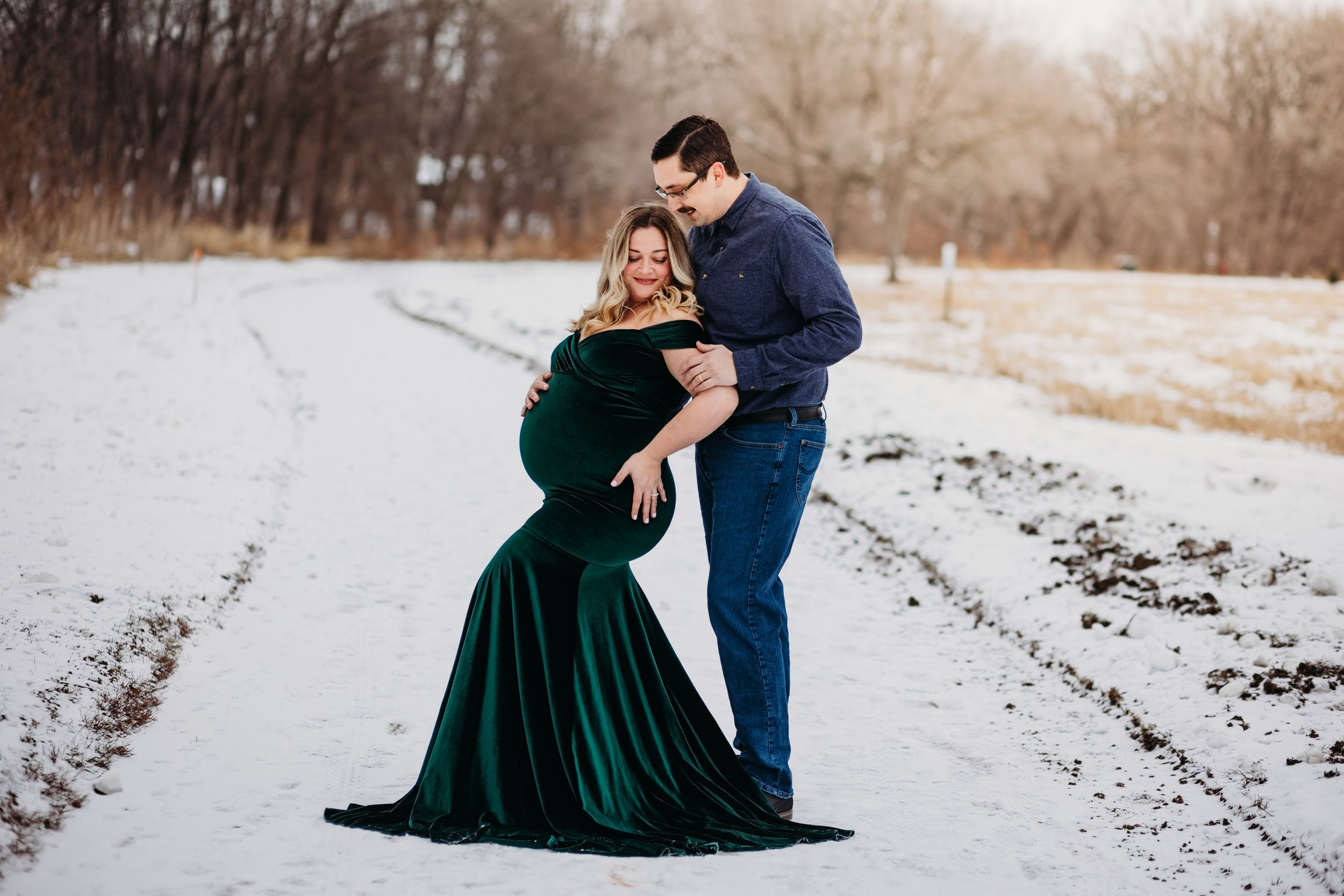 A couple embraces on a snowy path, with the woman wearing an elegant emerald velvet gown against a winter landscape.