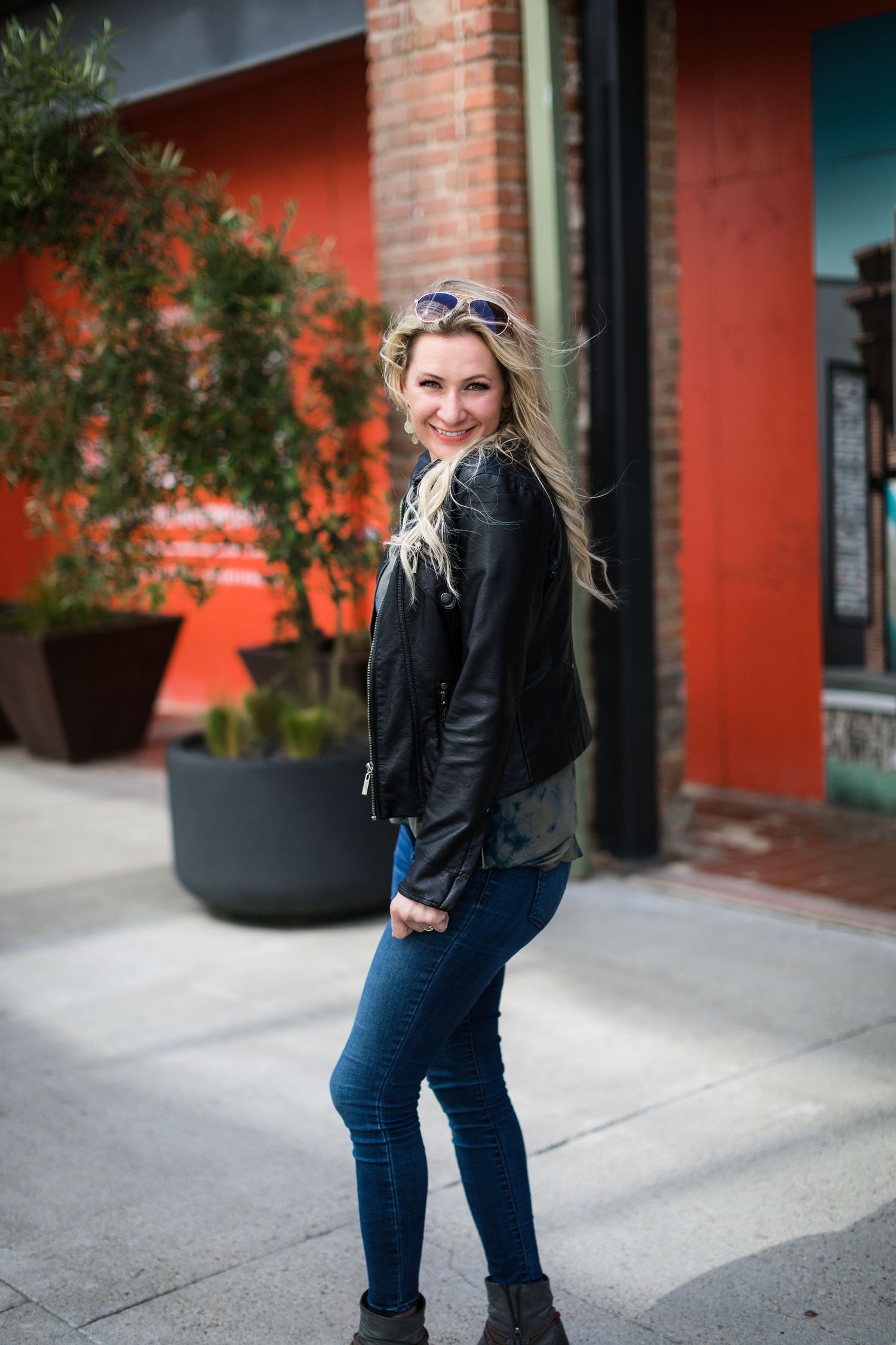 A woman wearing a black leather jacket and jeans stands smiling in front of a red wall with potted plants.