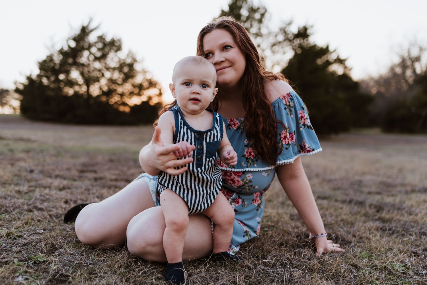 A parent and child sit together on dry grass at sunset.