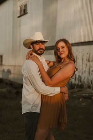 Couple in western attire poses at sunset against rustic barn wall.