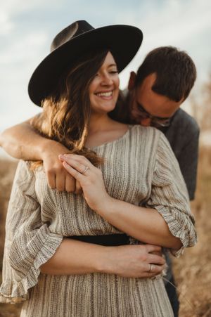 A candid engagement photo showing a couple embracing outdoors with the woman wearing a knit sweater and wide-brim hat.