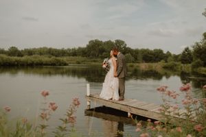 Wedding couple embracing on wooden dock over pond with wildflowers in foreground.