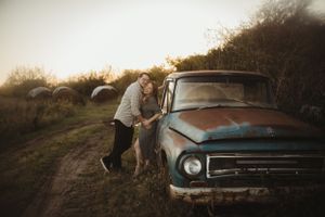 Couple poses playfully against vintage rustic truck in countryside setting.