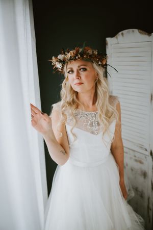 Bride in white lace dress and floral crown poses by window in romantic bridal portrait.