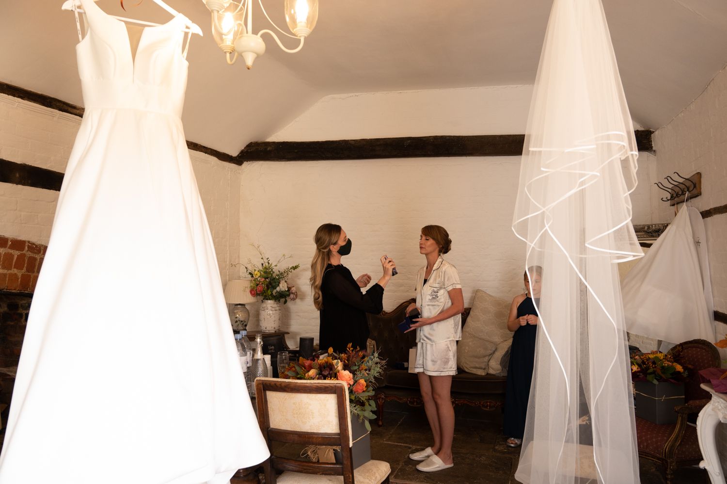 Wedding dress hanging in preparation room with people getting ready in background.
