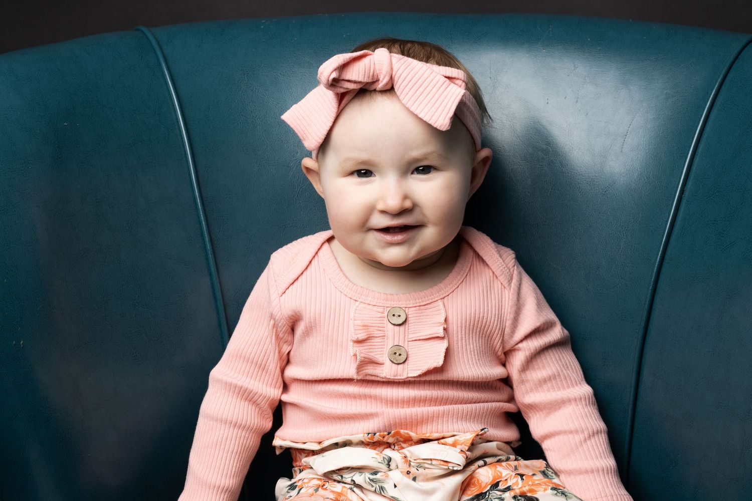 Baby wearing pink bow headband and cardigan sitting on blue leather chair.
