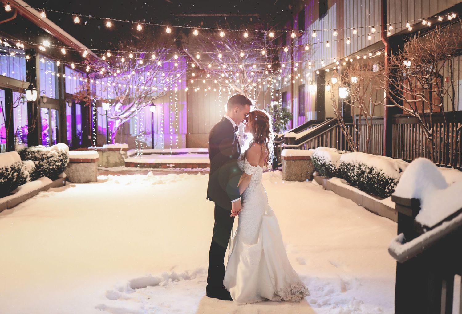Romantic winter wedding scene with couple embracing under string lights and snow.