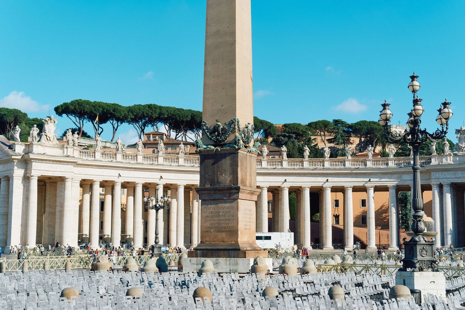 View of Saint Peter's Square colonnade with tall obelisk and Mediterranean pine trees.