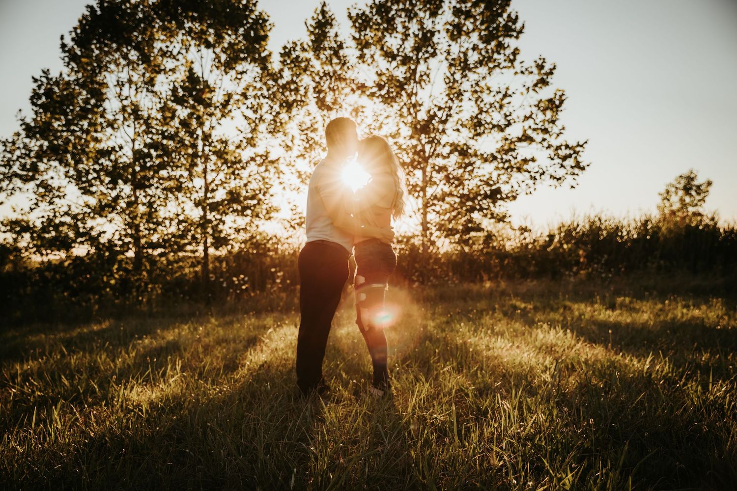 Silhouette of people embracing in a field with sun rays streaming through trees.