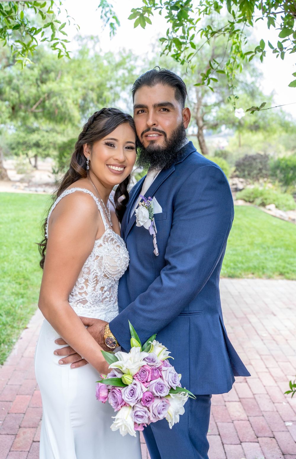 A couple poses for wedding photos on a brick pathway surrounded by lush greenery and holding a purple bouquet.