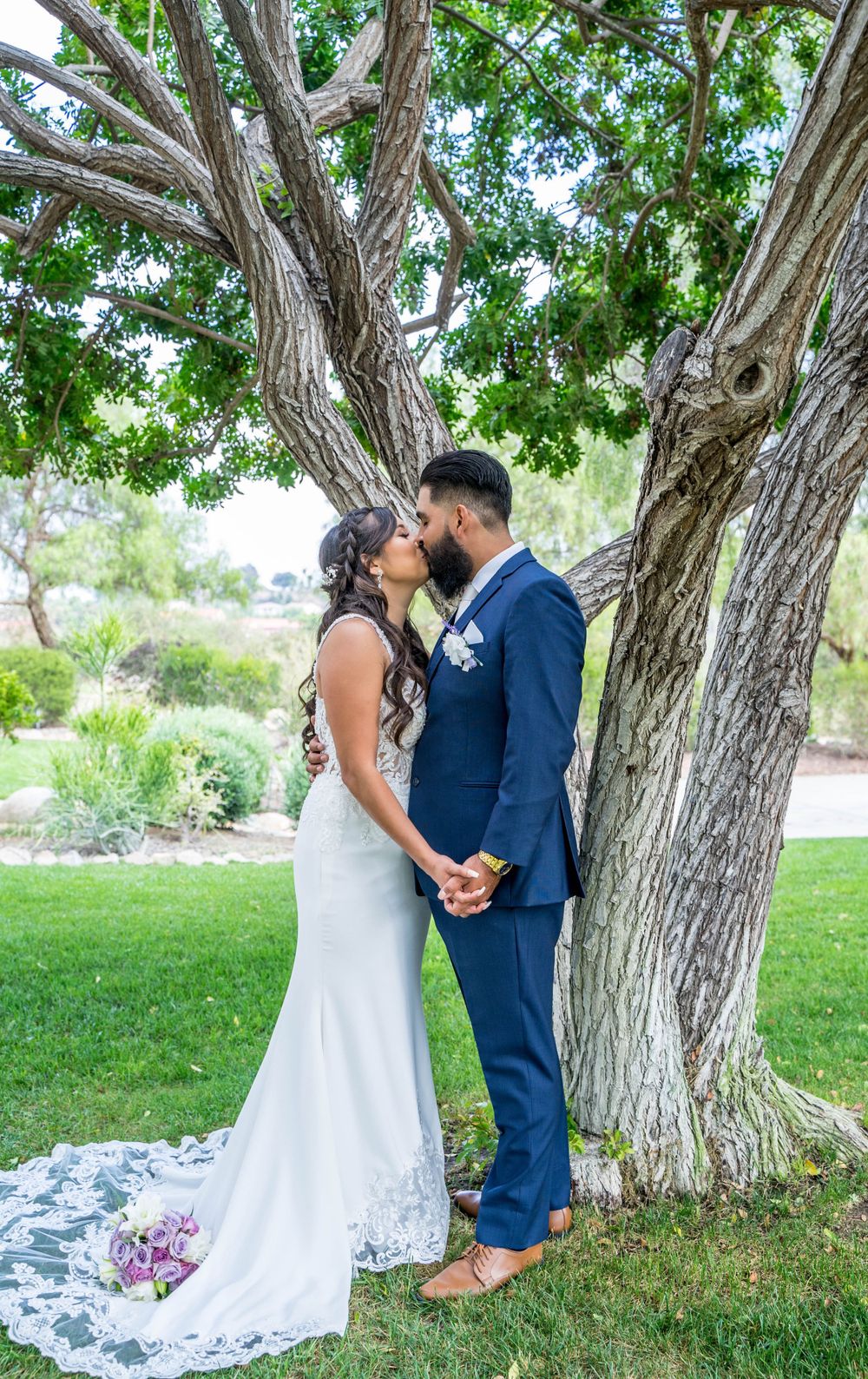 A romantic wedding couple embraces under trees in a garden setting.