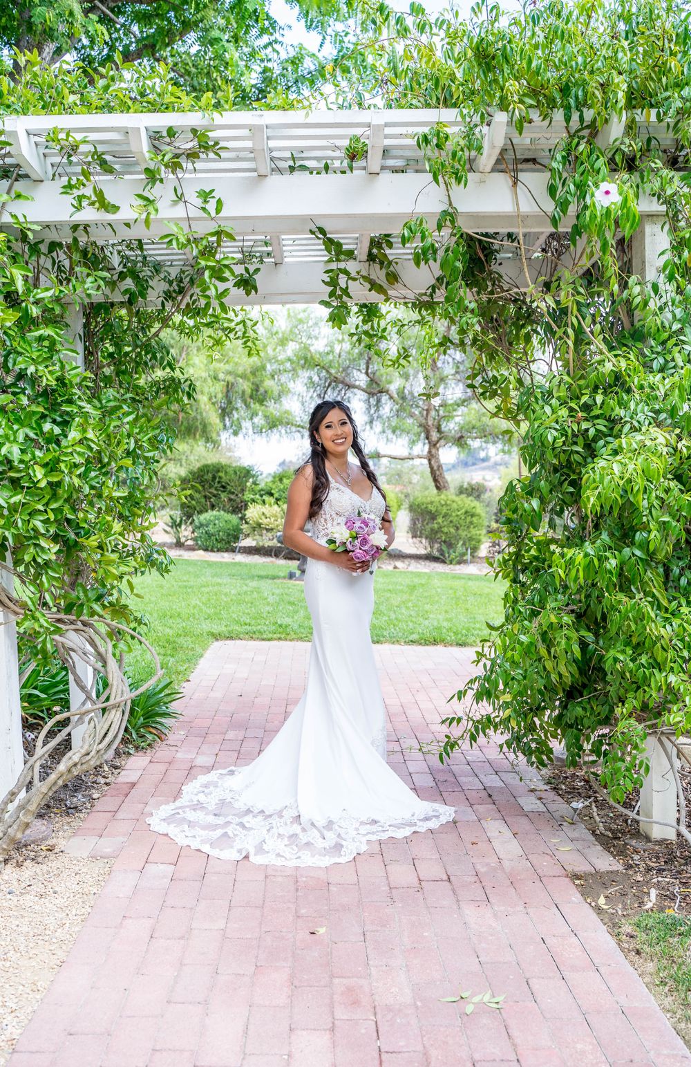 A bride in a white lace gown poses under a garden pergola with brick pathways and greenery.