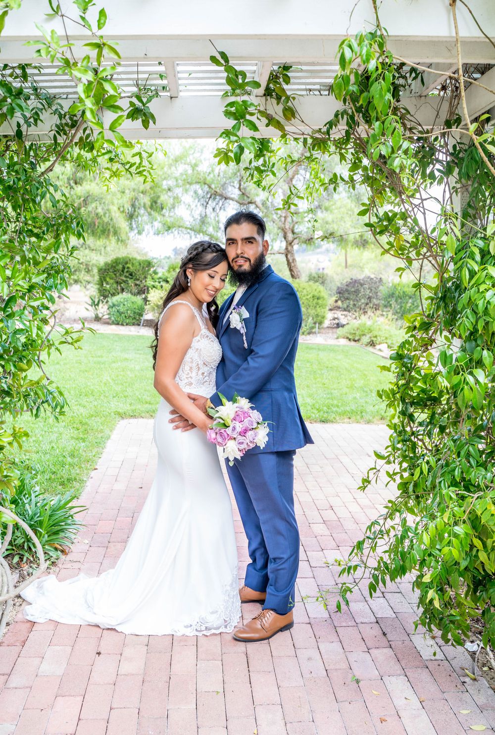 A couple in wedding attire shares romantic moments under green vines on a brick pathway in an outdoor garden setting.