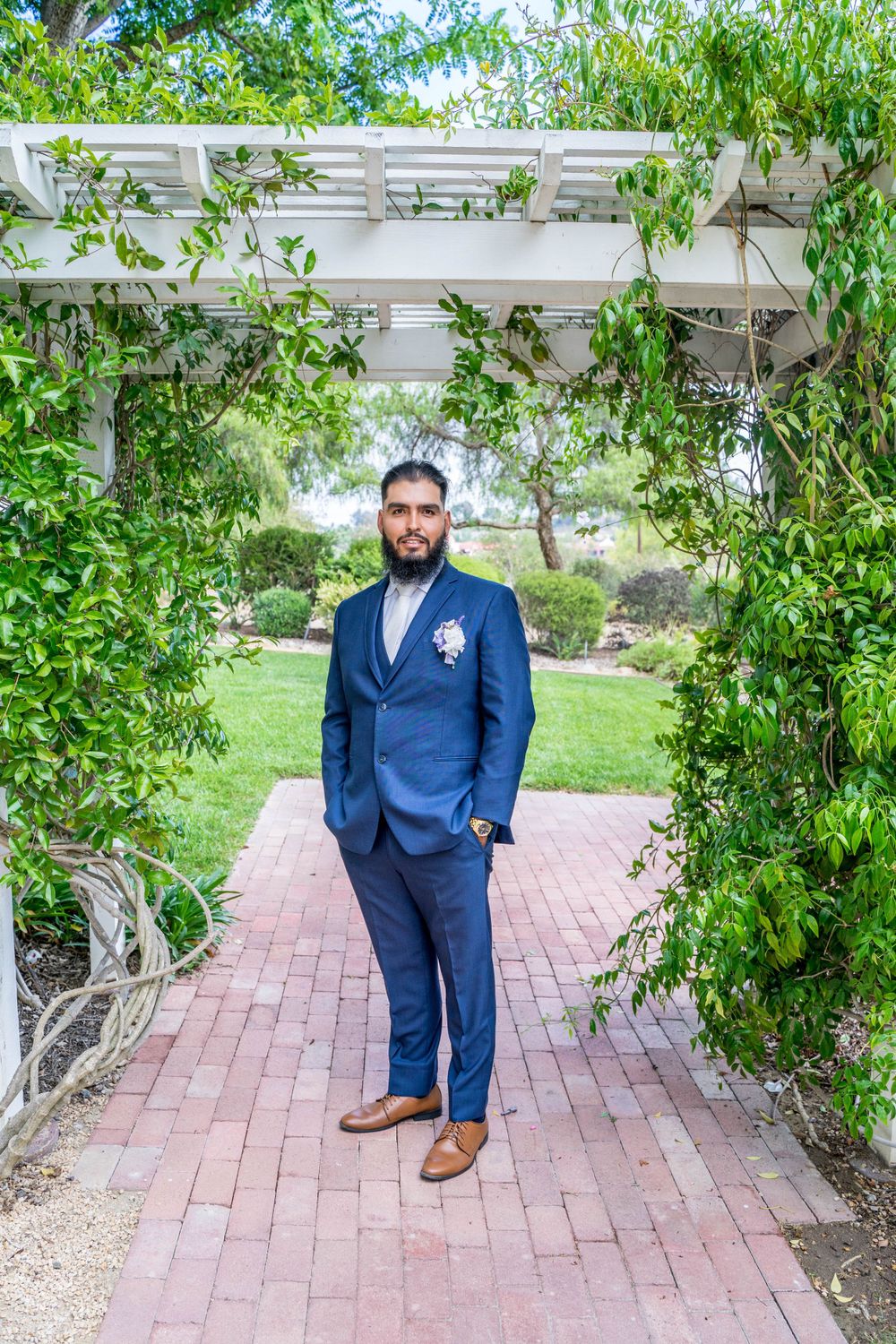 A groom in a navy blue suit stands alone on a brick pathway under a white pergola.