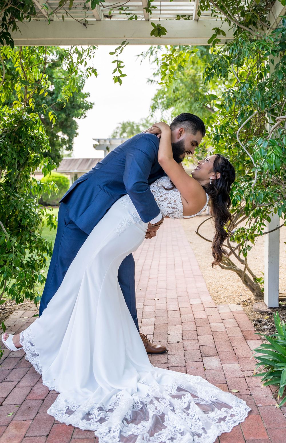 A wedding couple performs a romantic dip pose on a brick pathway under a pergola.