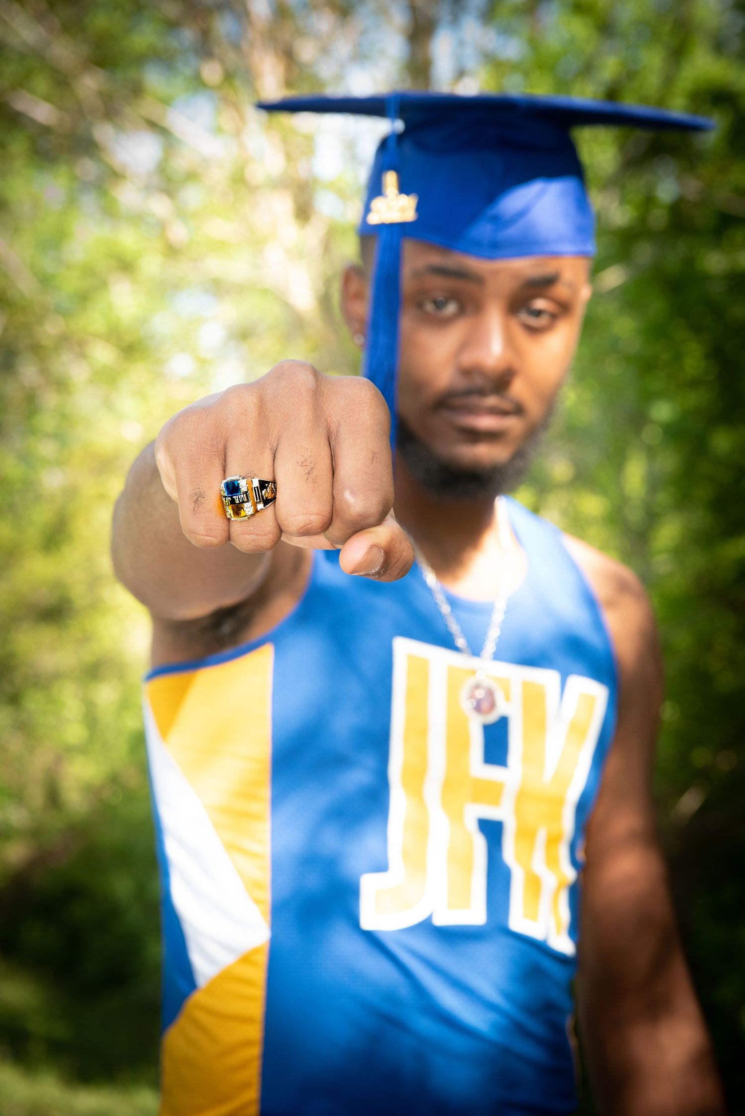 Graduate in blue and gold cap and gown shows off class ring in outdoor portrait session.