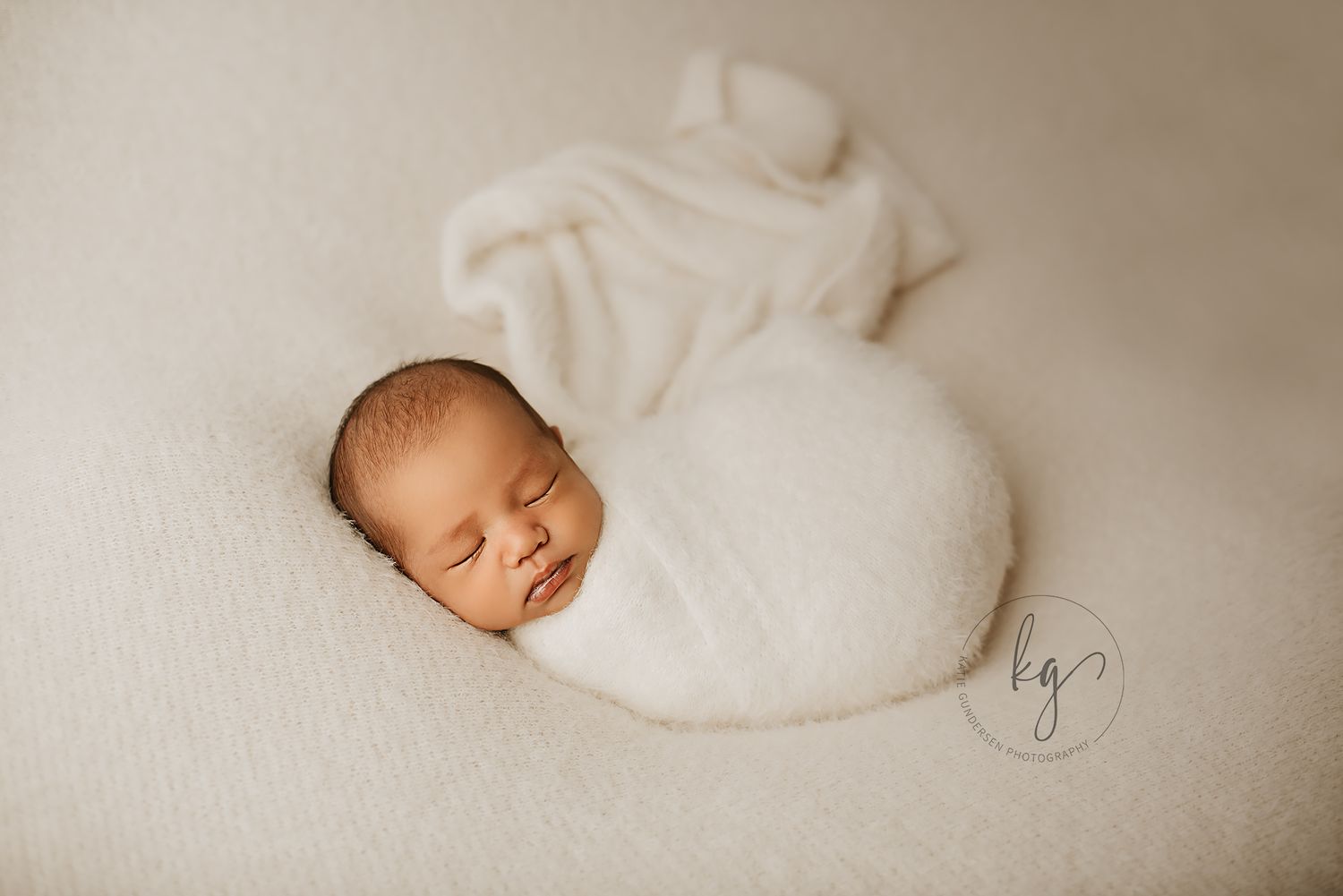 Newborn baby sleeps peacefully wrapped in white fabric against a neutral background.