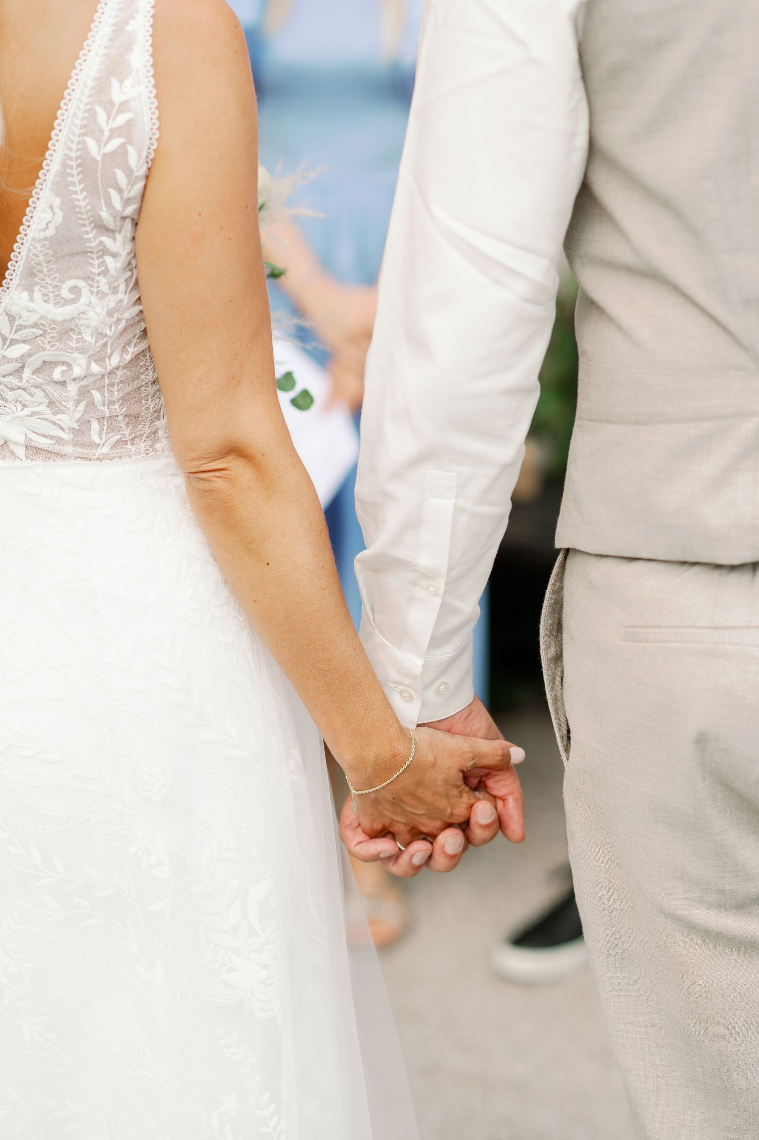 Close-up of newlyweds holding hands, showing white dress and light gray suit details.