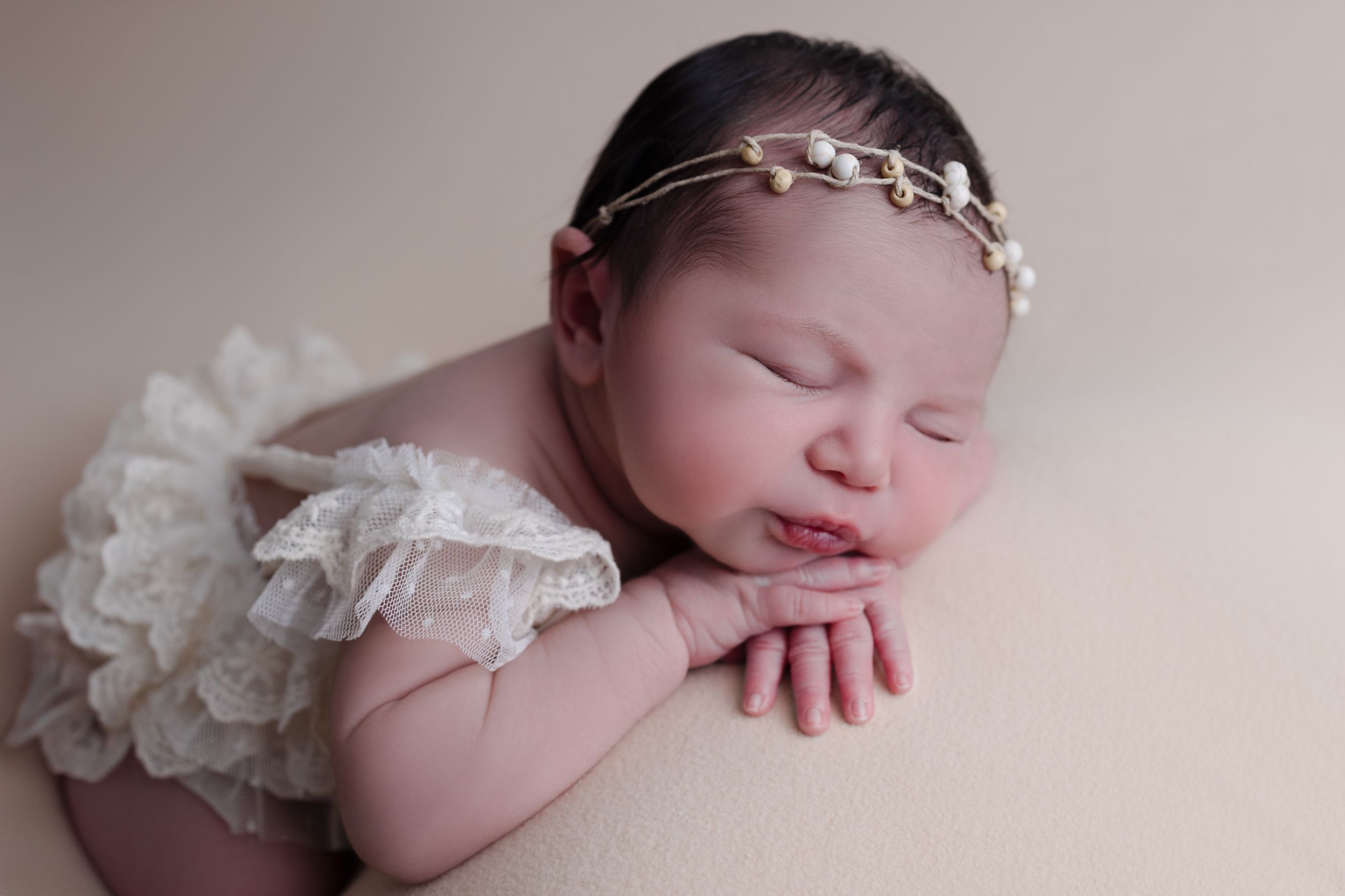 Sleeping newborn baby in cream lace outfit and delicate headband rests peacefully with hands folded.