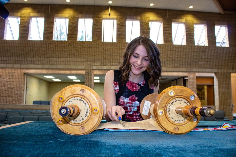 A woman examining Torah scrolls on a blue-covered table under window lighting.