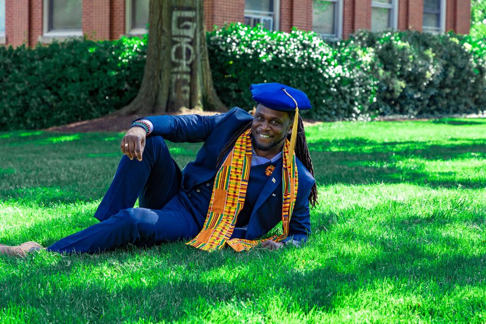 A graduate in blue regalia and gold stole relaxes on green lawn during graduation photos.
