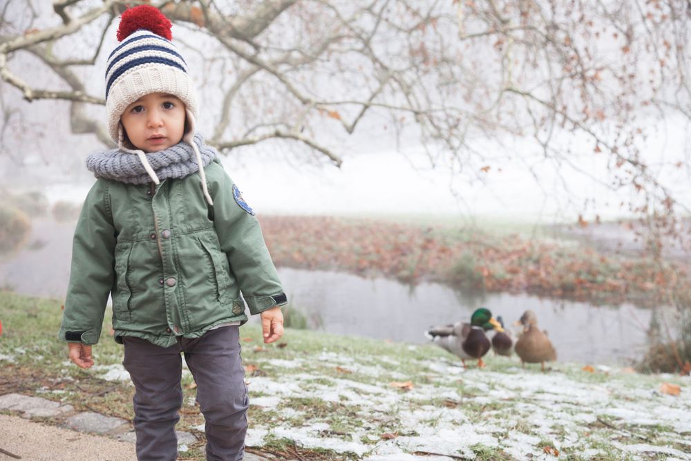 Portrait d'un jeune enfant dans un parc. Il porte un bonnet sur la tête et il y a des canards en arrière-plan.
