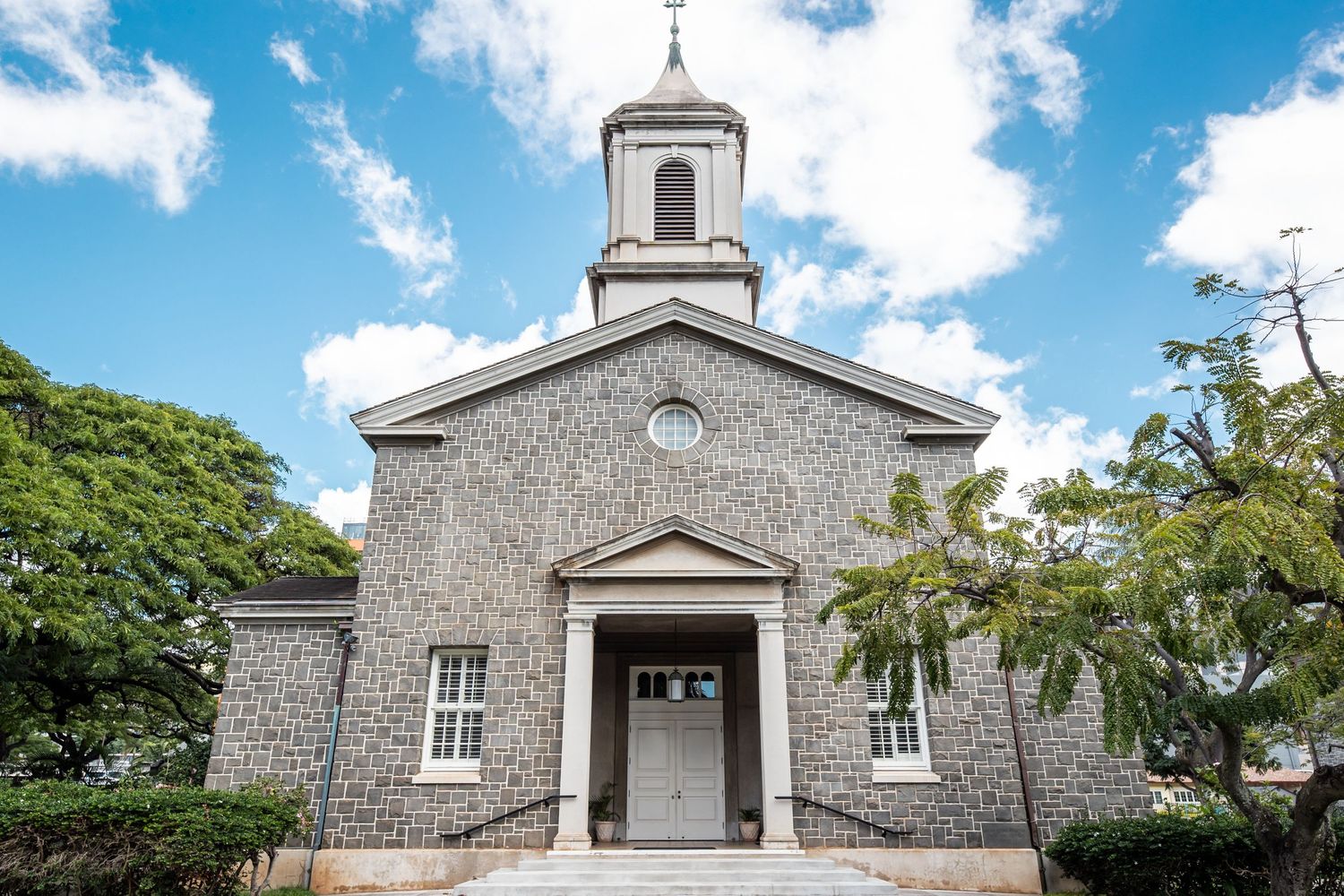 Historic coral stone church with bell tower, white columns, and steps under blue sky in Hawaii.