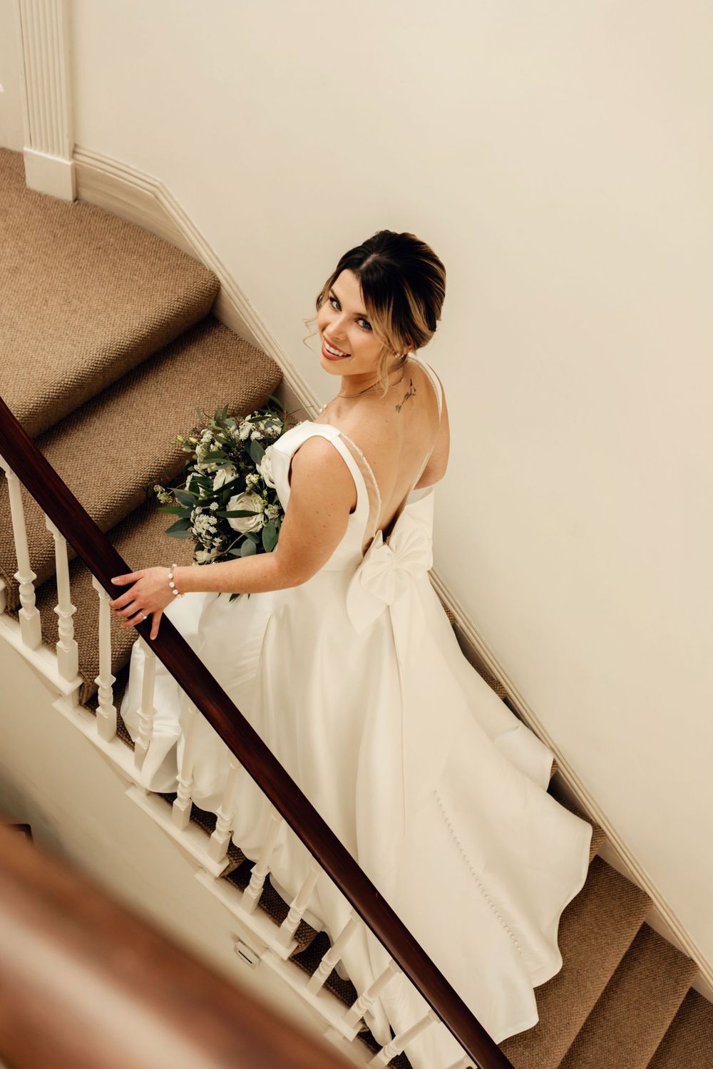 Wedding gown photographed from behind on carpeted staircase showing bow detail and train.