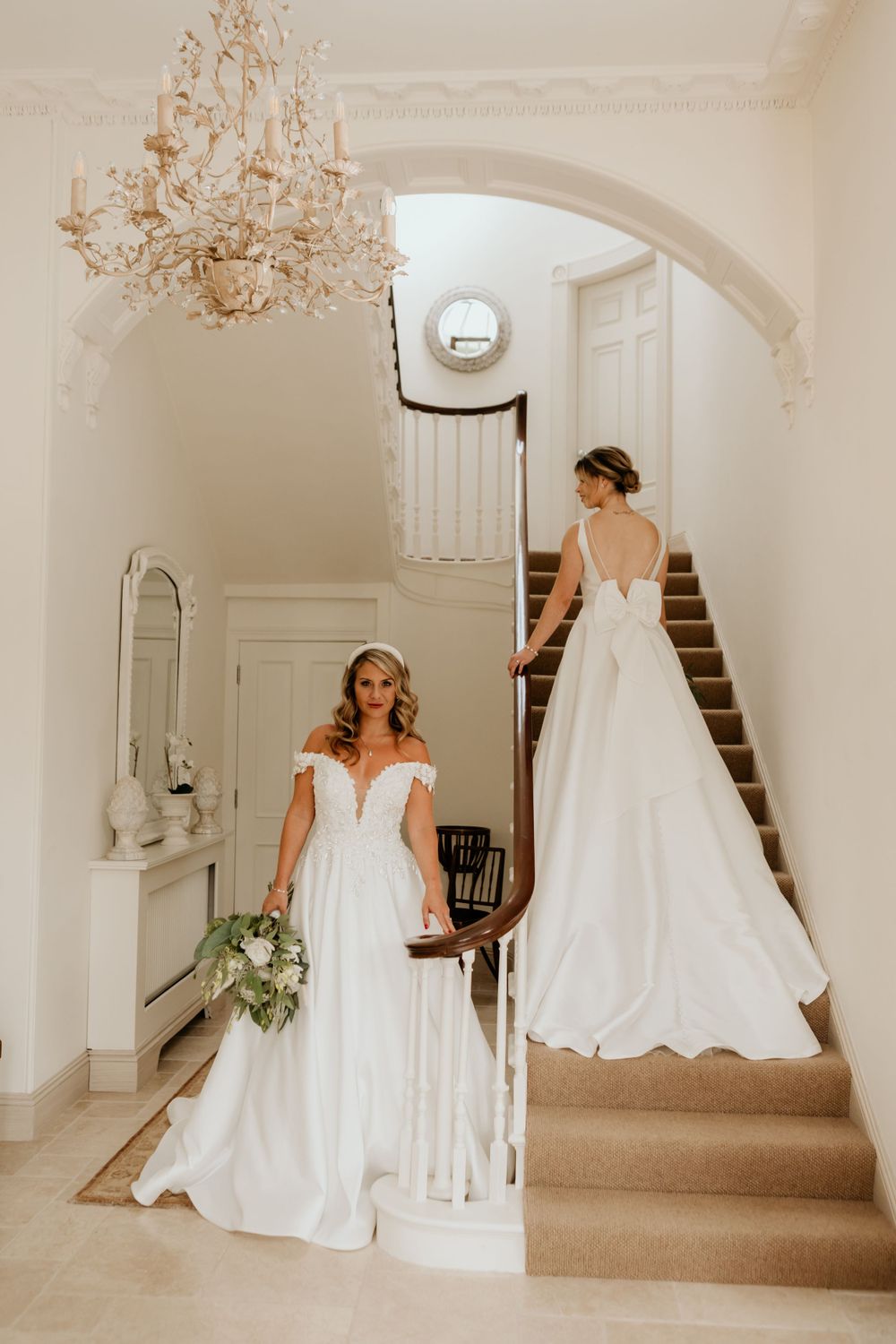 Two wedding gowns showcased on elegant staircase with crystal chandelier and classic interior details.