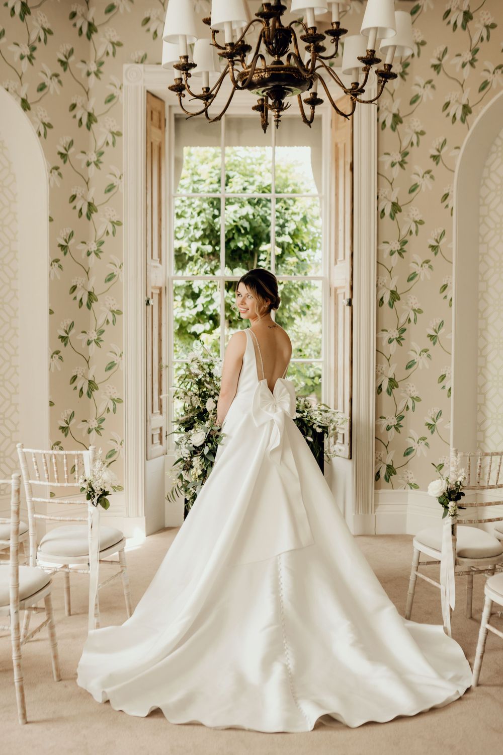 Wedding gown with dramatic train and bow detail photographed in elegant room with floral wallpaper and chandelier.