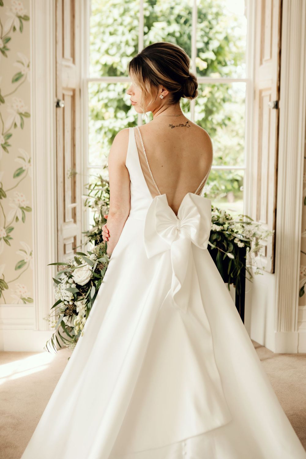 Elegant wedding dress with deep V-back and dramatic bow detail shown in sunlit room with floral wallpaper.