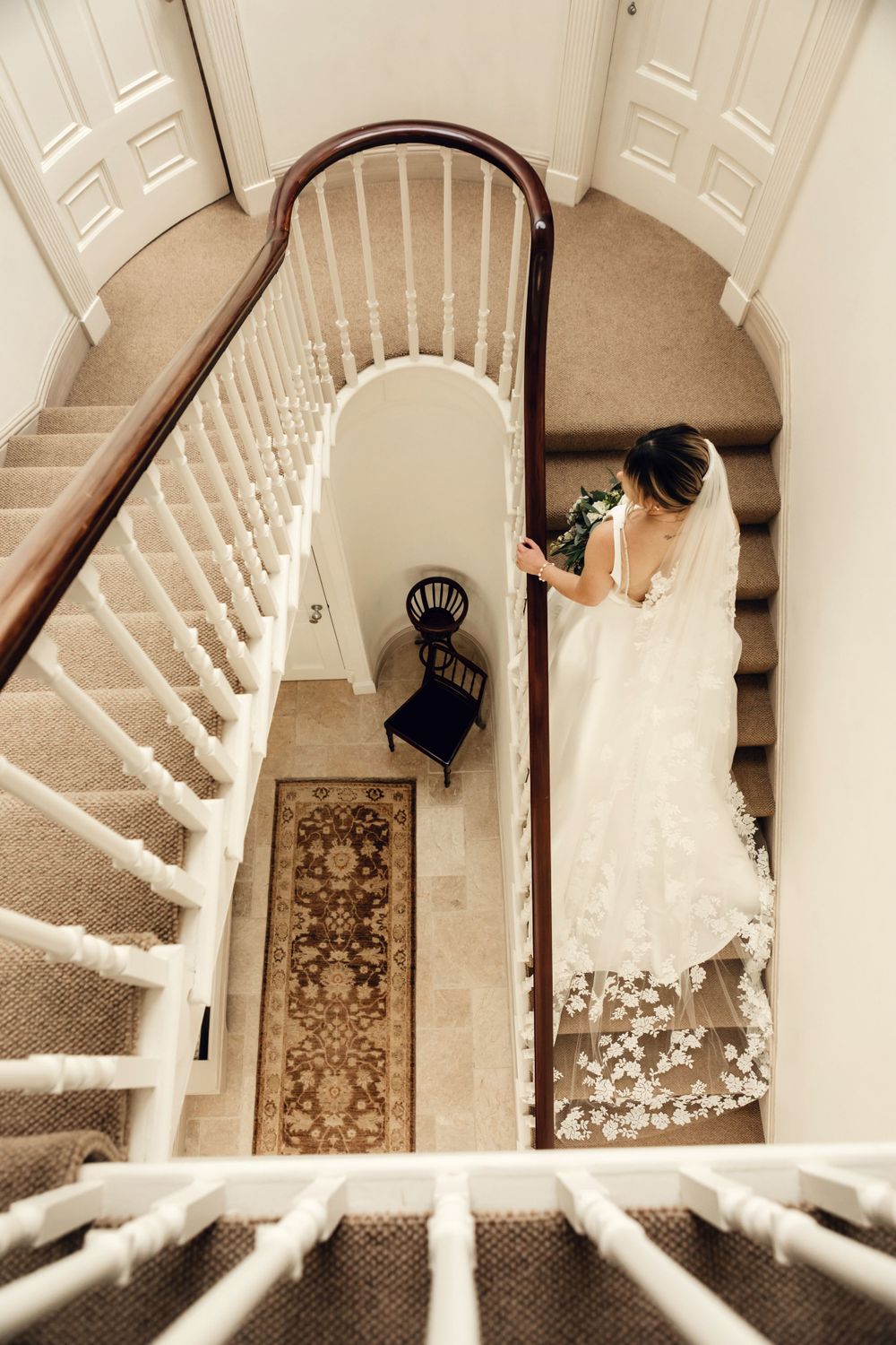 Aerial view of an elegant staircase with white spindles and a person in a white gown descending the carpeted steps.