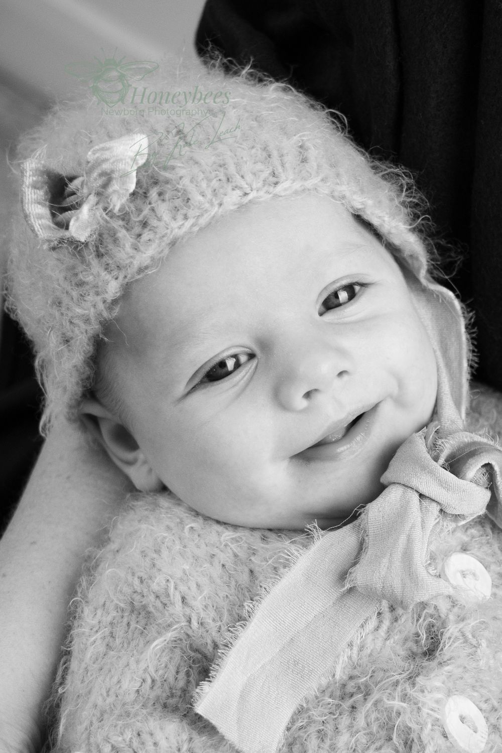 smiting baby boy with knitted Bonet with flower on tied under his chin. mums arm is in the background