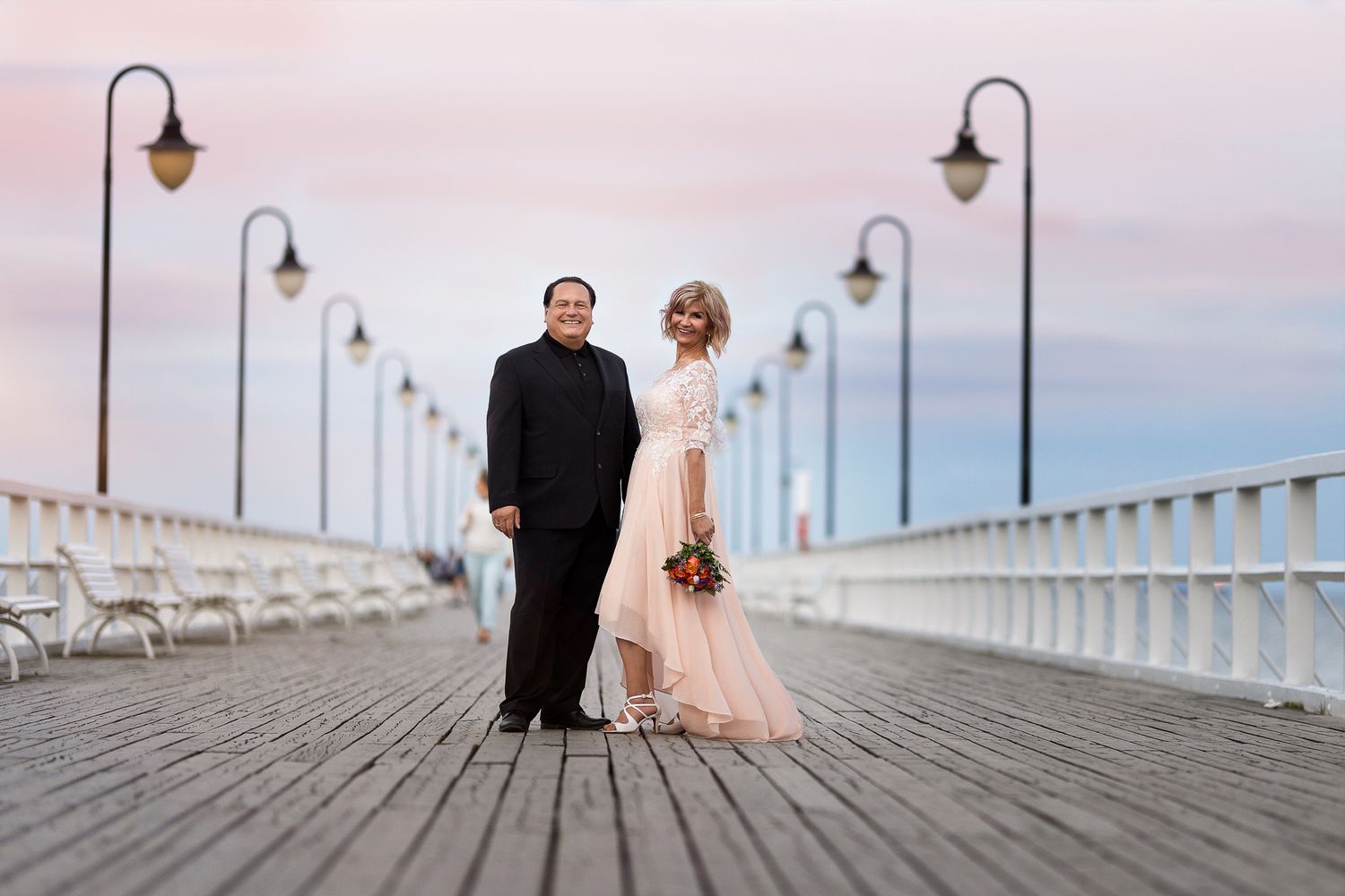 Wedding couple walking along a wooden pier with vintage-style lampposts at sunset.