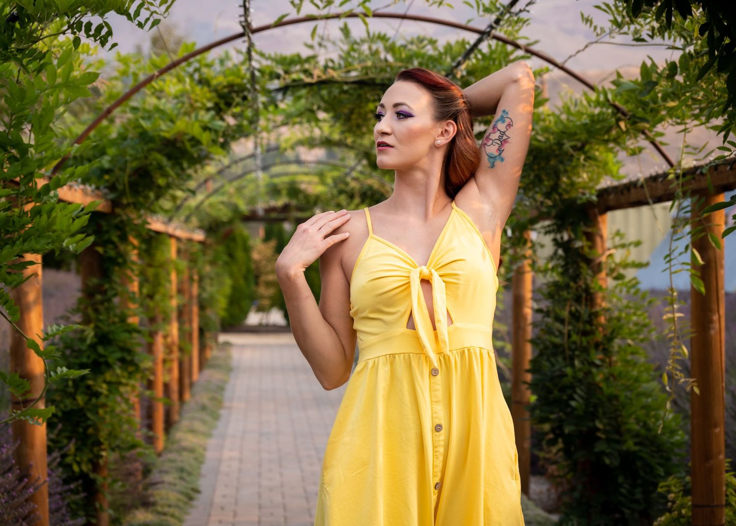 Redhead woman in a yellow sundress poses under a wooden garden arch surrounded by greenery.