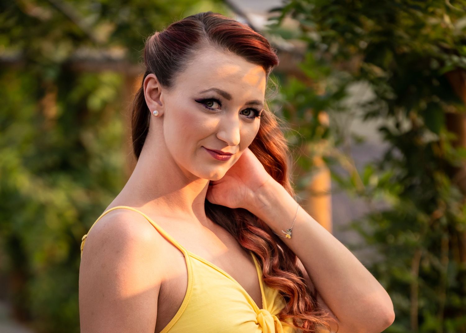 Smiling redhead woman in a yellow dress poses outdoors with lush green foliage in the background.