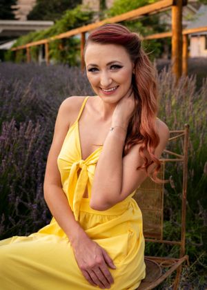 Smiling redhead woman in a yellow sundress sits in a lavender field on a vintage chair.