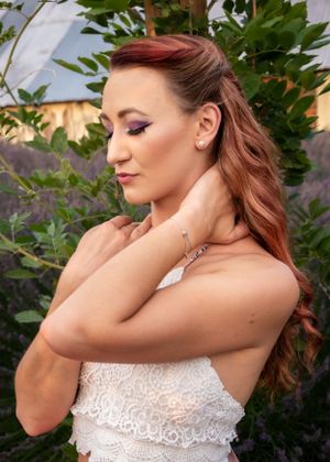 Redhead woman in a white lace top poses among green foliage with purple eye makeup and a bracelet.
