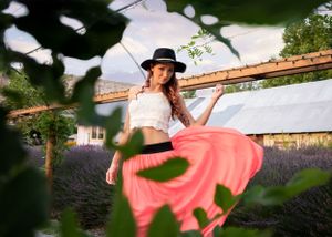 Woman in a white crop top and flowing pink skirt twirls in a lavender garden wearing a black hat.